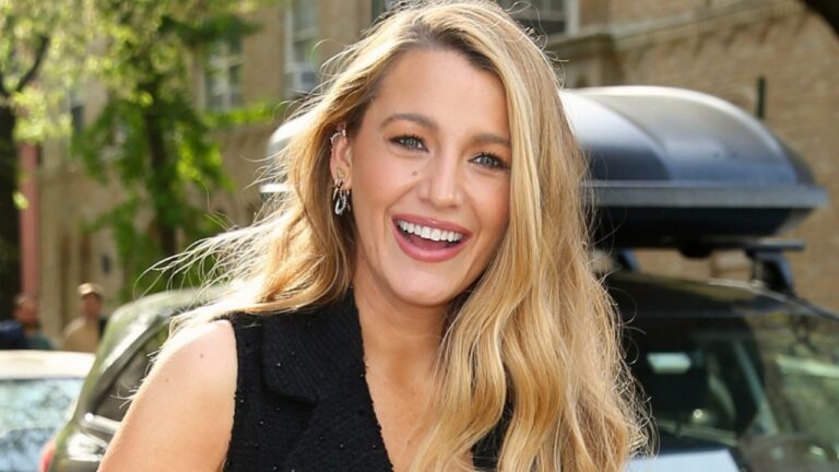 Mulher sorri ao ar livre em uma rua ensolarada, com carros e prédios ao fundo, vestindo roupa preta sem mangas e usando brincos prateados