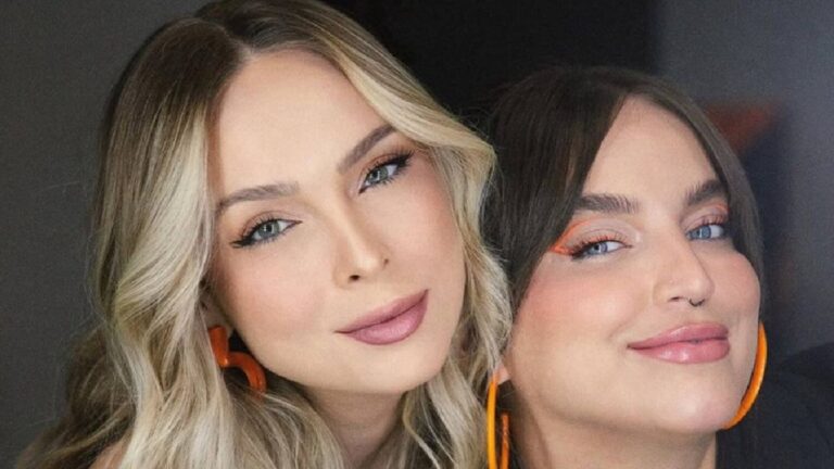 A imagem mostra duas mulheres posando juntas e sorrindo. Ambas estão maquiadas, com destaque para o delineado e os brincos laranja combinando. A mulher à esquerda tem cabelo loiro e ondulado, enquanto a da direita tem cabelo escuro e liso com franja. O fundo é neutro, destacando os rostos e os acessórios das duas