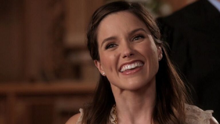 Mulher sorri amplamente em ambiente interno com iluminação quente. Ela tem cabelos castanhos penteados para trás, usa brincos discretos e uma blusa clara, transmitindo alegria e simpatia