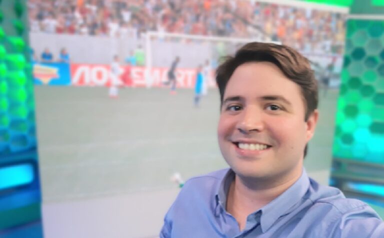 Homem sorri enquanto tira uma selfie em um estúdio de TV com temática esportiva, tendo ao fundo uma tela que exibe uma partida de futebol e arquibancada com torcedores