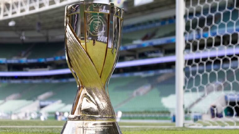 Taça dourada do Campeonato Paulista posicionada no gramado de um estádio de futebol, próxima à trave, com arquibancadas desfocadas ao fundo