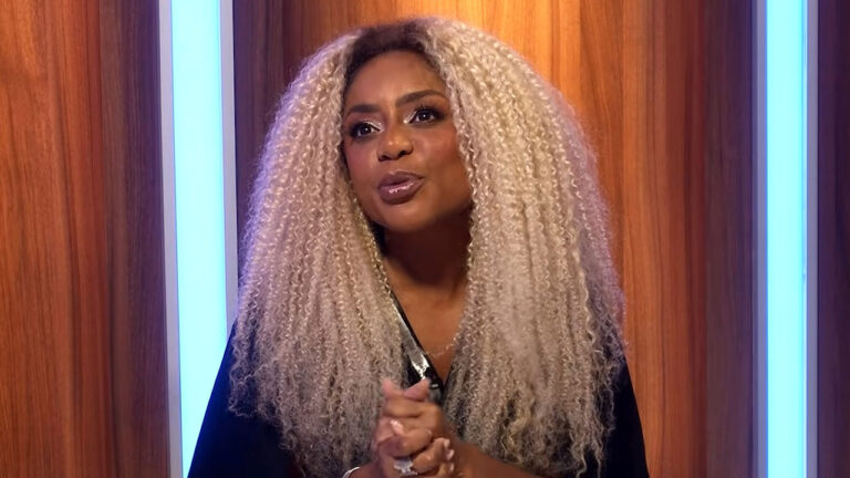 Cariúcha, com cabelos loiros cacheados volumosos, em close-up, vestindo preto, em um cenário com painéis de madeira e luzes azuis