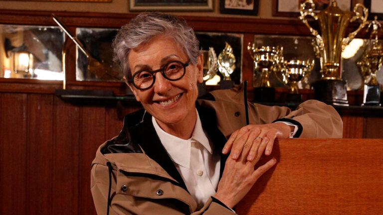 Cássia Kis com cabelo grisalho curto e óculos redondos sorri em ambiente com troféus ao fundo