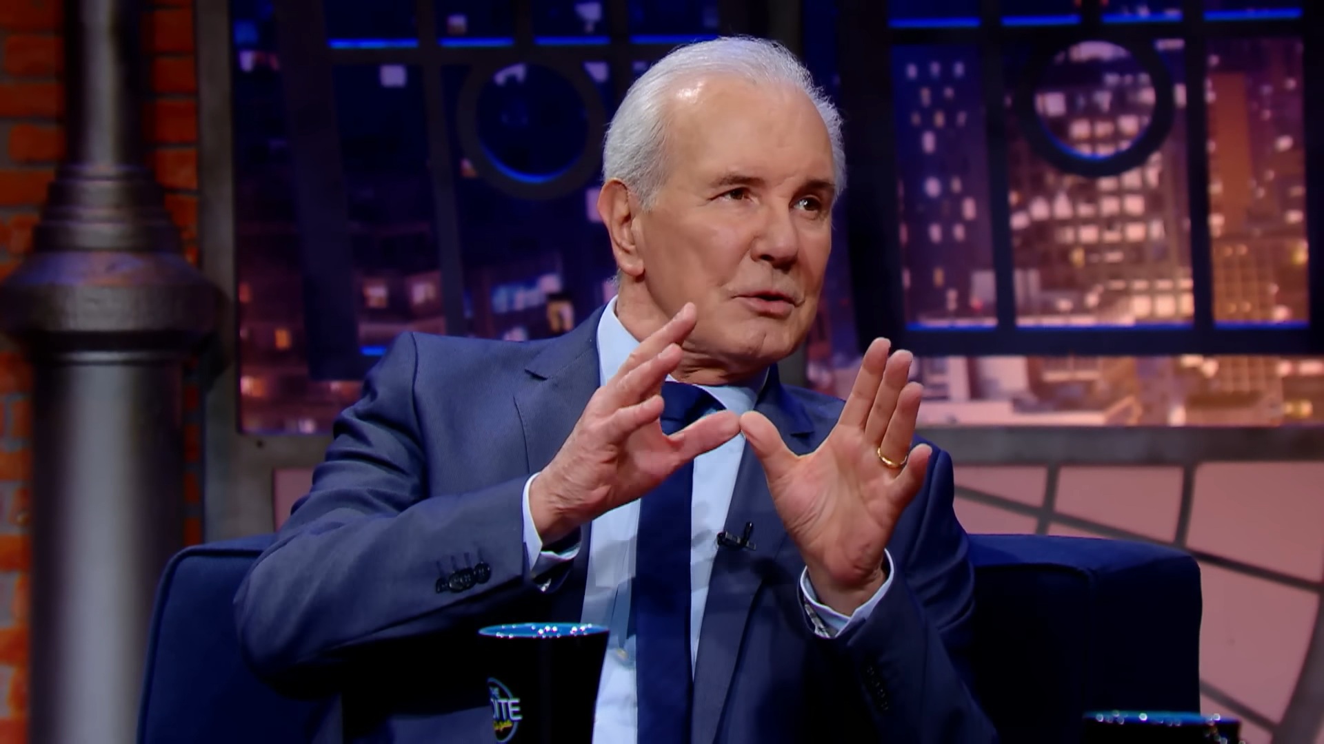 A imagem mostra Celso Freitas, um homem de cabelos grisalhos e terno azul participando de uma entrevista no programa The Noite. Ele gesticula com as mãos enquanto fala, demonstrando envolvimento na conversa. Ao fundo, há um cenário com luzes que simulam prédios noturnos e uma caneca preta com o logotipo do The Noite sobre a mesa