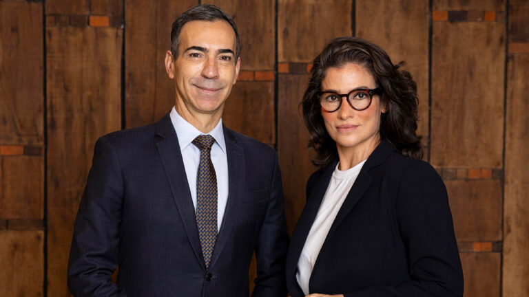 César Tralli de terno e Renata Vasconcellos de óculos com blazer preto posam juntos em estúdio de fundo amadeirado