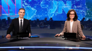 César Tralli de terno e Renata Vasconcellos de blazer marrom sorrindo na bancada do Jornal Nacional da Globo