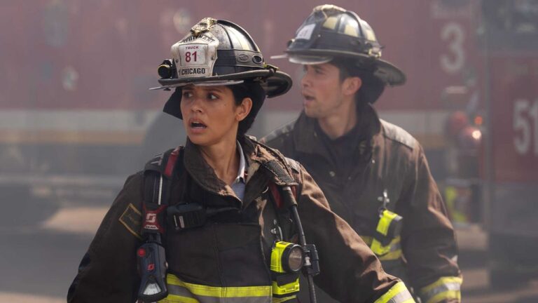 Na série Chicago Fire, dois bombeiros em ação aparecem em meio à fumaça, usando uniforme de proteção completo e capacetes identificados com Truck 81 – Chicago. A bombeira em primeiro plano olha para o lado com expressão de alerta, enquanto o colega ao fundo observa a cena, sugerindo uma situação de emergência em andamento