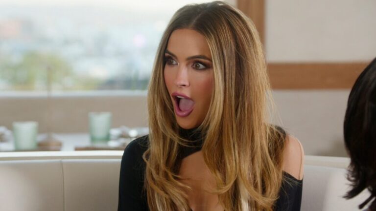 Mulher de cabelos longos e loiros, vestida de preto, aparece com expressão surpresa e boca aberta em ambiente interno iluminado