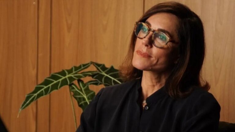 Christiane Torloni usando óculos de armação grossa, cabelo castanho na altura dos ombros e expressão serena. Ela veste uma blusa escura e aparece sentada ou inclinada levemente, como se estivesse ouvindo alguém com atenção