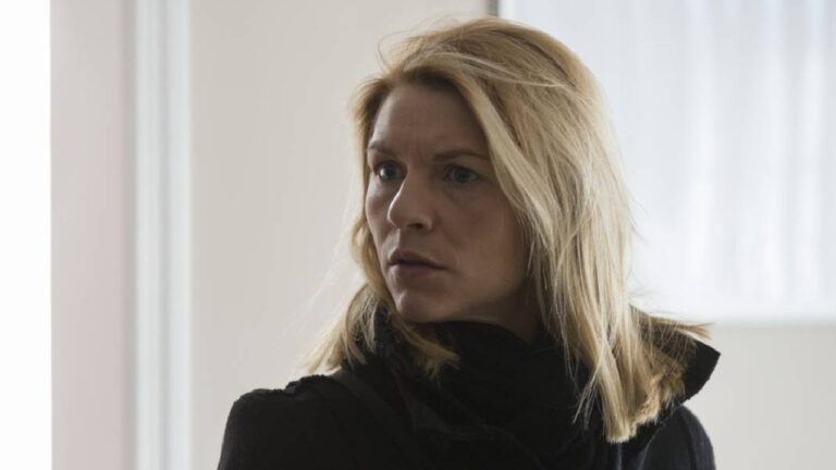 Mulher loira de expressão séria e olhar atento, vestindo casaco escuro, está em um ambiente iluminado por luz natural. A cena transmite tensão e apreensão, como se ela tivesse acabado de receber uma notícia preocupante ou percebido algo inesperado