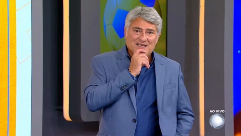 Homem de cabelos grisalhos apresenta programa de TV ao vivo, vestindo terno azul e sorrindo, com cenário colorido e uma bola de futebol exibida em tela ao fundo