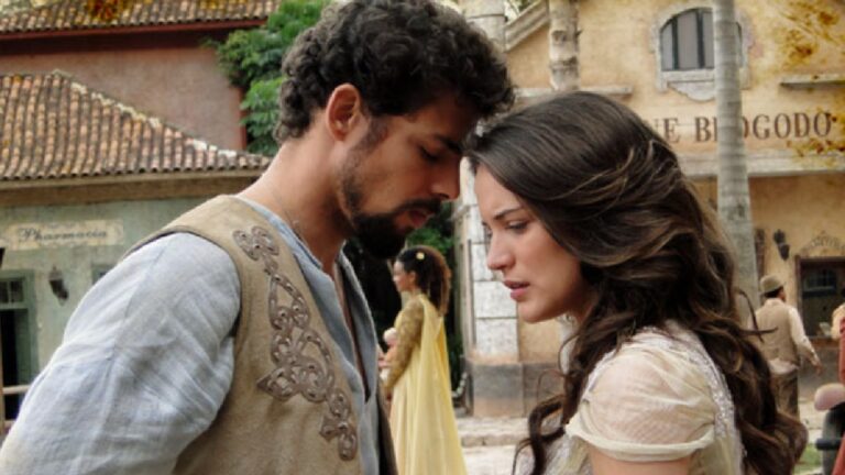 Cena de época mostra um homem e uma mulher encostando as testas um no outro em um momento de emoção. Ambos têm expressões sérias e estão vestidos com roupas antigas, em um cenário que remete ao interior colonial