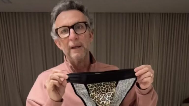 A imagem mostra um homem usando óculos e um suéter rosa, segurando uma peça de roupa íntima com estampa de onça. Ele está em um ambiente interno, com cortinas fechadas ao fundo, e parece estar falando ou explicando algo enquanto exibe o item diante da câmera