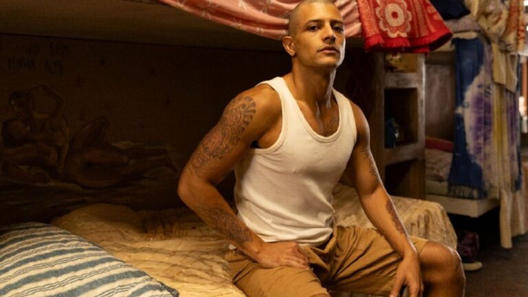 Homem de cabeça raspada e tatuagens nos braços está sentado em uma cama, vestindo regata branca e bermuda, em um ambiente simples que parece um quarto coletivo