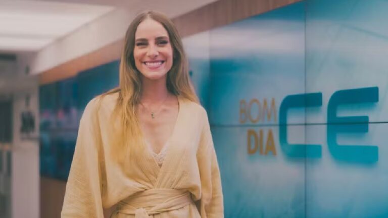 Uma mulher sorri diante de um painel luminoso que exibe o título “Bom Dia CE”. Ela tem cabelos longos e loiros, veste um robe bege cinturado e usa um colar delicado. A cena ocorre em um estúdio de televisão moderno, com iluminação suave e corredores ao fundo, transmitindo um clima acolhedor e profissional