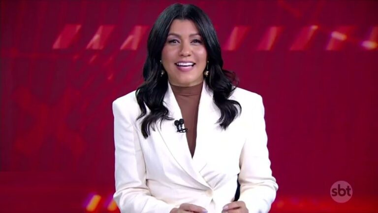 Daniele Brandi, que é uma mulher de cabelos pretos e ondulados, enquanto apresenta o programa Aqui Agora no estúdio do SBT, vestindo blazer branco sobre blusa marrom, com microfone preso à roupa e fundo vermelho iluminado com o logotipo da emissora no canto da tela