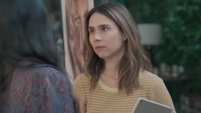 A imagem mostra uma mulher de cabelos castanhos e expressão séria, segurando um tablet. Ela veste uma blusa amarela clara com textura e está de frente para outra pessoa, que aparece de costas