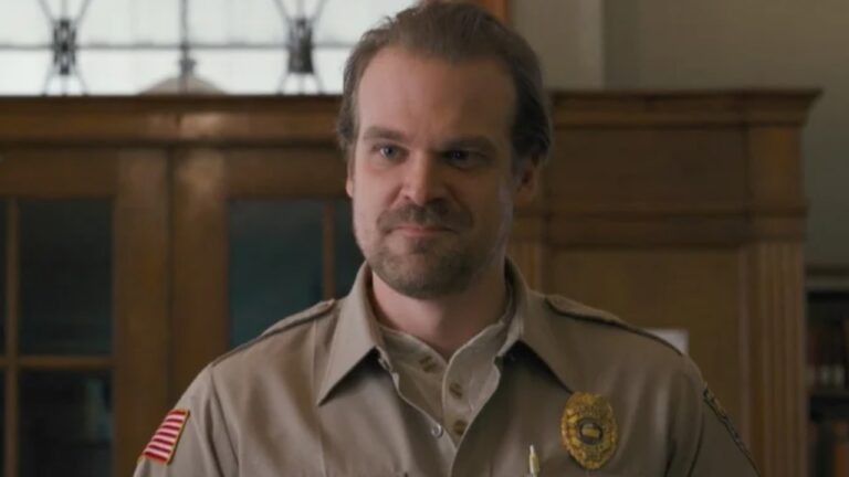 A imagem mostra um homem usando um uniforme bege de xerife, com um distintivo dourado e uma bandeira dos Estados Unidos no ombro. Ele tem expressão séria, barba curta e cabelo penteado para trás. O cenário ao fundo parece ser o interior de um prédio público, com portas e painéis de madeira