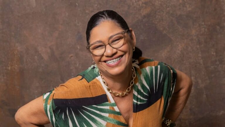 Mulher sorridente posa diante de fundo neutro, usando óculos, colar dourado e blusa colorida com estampa tropical em tons de verde e laranja