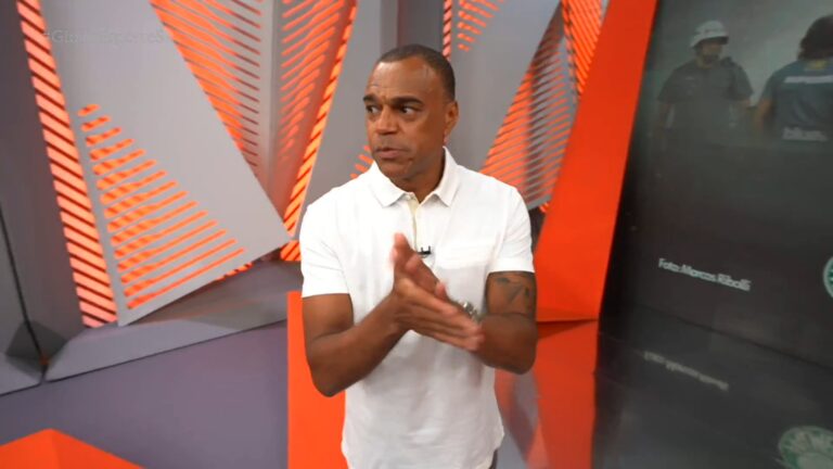 Denílson veste camisa polo branca e aparece em estúdio moderno com painéis nas cores cinza e laranja, em ambiente que lembra um programa esportivo de televisão
