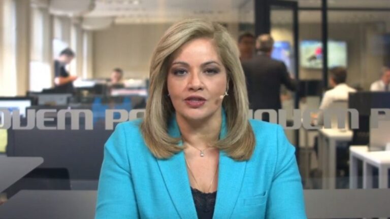 Denise Campos de Toledo com blazer azul fala em frente às câmeras, usando microfone de lapela, em redação de jornal com pessoas e computadores ao fundo