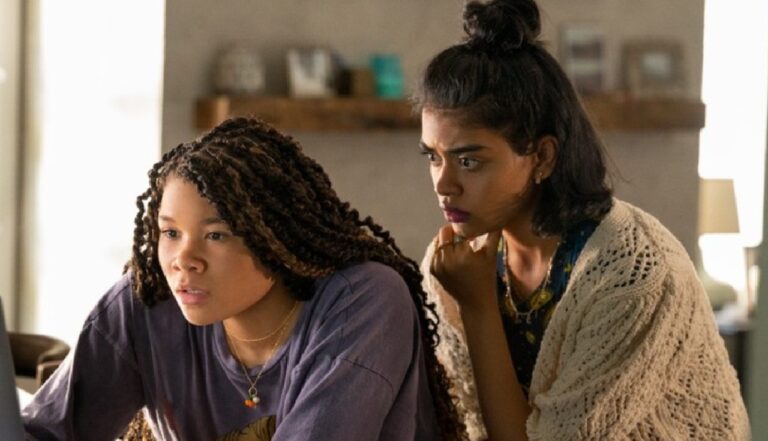 A imagem mostra duas mulheres concentradas olhando para algo em frente a elas, possivelmente uma tela de computador. A mulher à esquerda tem tranças longas e veste uma camiseta roxa, enquanto a mulher à direita tem o cabelo preso em um coque e está usando um cardigã de crochê claro. Ambas parecem preocupadas ou intrigadas com o que estão vendo, em um ambiente doméstico iluminado pela luz natural