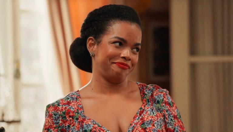 A imagem mostra uma mulher com expressão levemente divertida ou irônica, vestindo um vestido floral colorido e batom vermelho. Ela está em um ambiente interno bem iluminado, que parece ser uma sala decorada