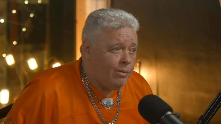 DJ Marlboro usando camisa laranja e colares prateados falando diante de microfone em estúdio de gravação iluminado