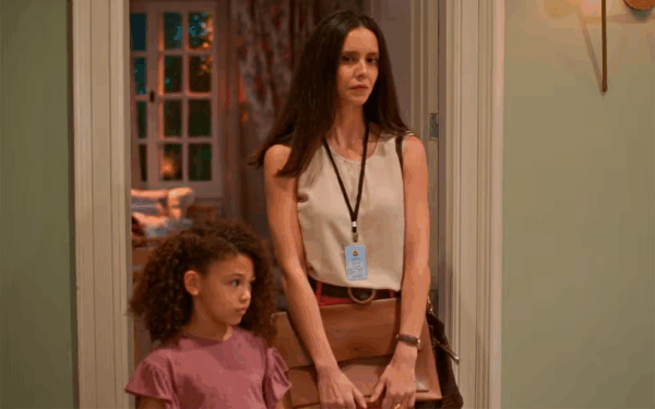 Mulher com expressão séria, vestindo blusa clara e crachá no pescoço, está ao lado de uma menina de camiseta rosa em uma porta de casa, observando algo no interior do cômodo iluminado.