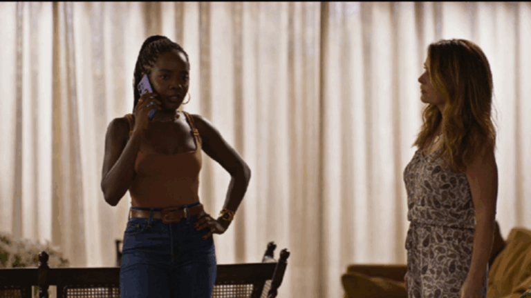 A imagem mostra duas mulheres em uma sala iluminada por luz suave, com cortinas claras ao fundo. À esquerda, uma mulher está ao telefone, com expressão séria, usando blusa marrom e calça jeans. À direita, outra mulher a observa atentamente, vestindo um macacão estampado