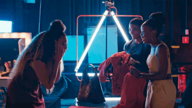 A imagem mostra três pessoas em um camarim ou estúdio, reunidas em volta de uma mesa com roupas e bolsas. Elas estão sorrindo e demonstrando empolgação, sugerindo um momento de celebração ou surpresa. O ambiente é iluminado por luzes de neon, criando um clima moderno e vibrante