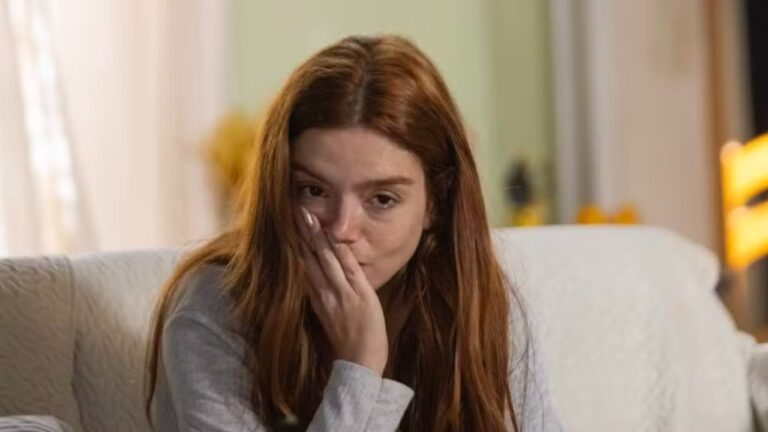 Jovem ruiva sentada no sofá com a mão sobre a boca em cena emocional dentro de sala iluminada