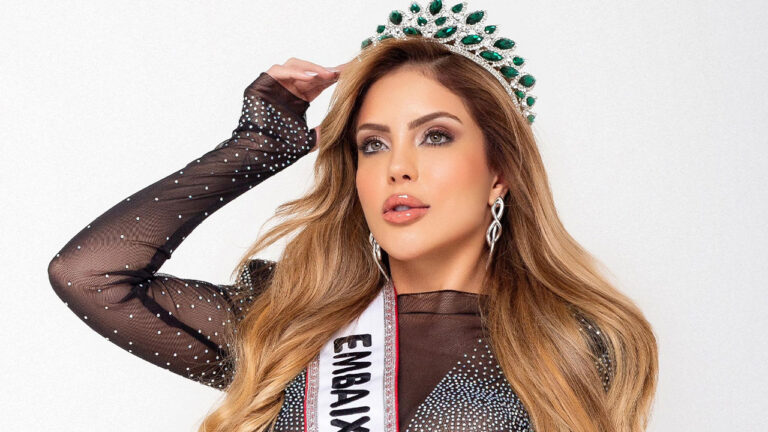 Duda Mattos posando com uma coroa de pedras verdes e faixa de Embaixatriz, usando vestido preto transparente com brilhos