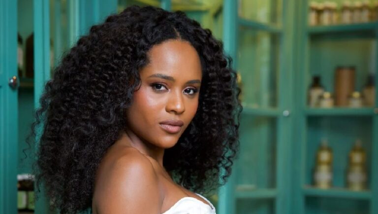 Mulher com cabelos cacheados e expressão confiante posa em ambiente interno com armários verde-água ao fundo, vestindo roupa clara e elegante