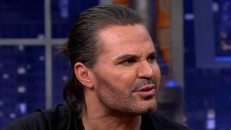 Homem de cabelo preso e barba rala fala com expressão séria em estúdio de programa noturno