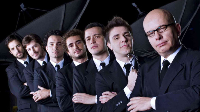 Elenco de apresentadores do CQC da Band em fila, de terno preto, braços cruzados, com fundo de antenas parabólicas