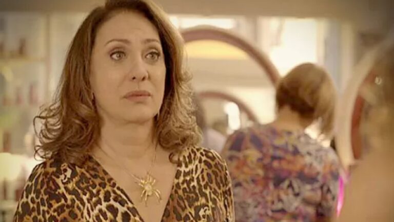 Mulher de expressão séria usa vestido com estampa de oncinha e colar dourado em formato de aranha, em um ambiente interno com espelhos e pessoas ao fundo, sugerindo um salão de beleza