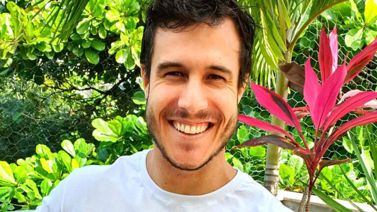 O ator Emiliano d'Ávila sorri com barba rala e camiseta branca em close-up em meio a uma vegetação com folhas verdes e vermelhas