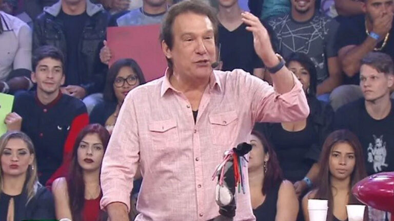 Emílio Surita com camisa rosa no palco do Pânico na Band, gesticula com a mão esquerda enquanto a plateia assiste ao fundo