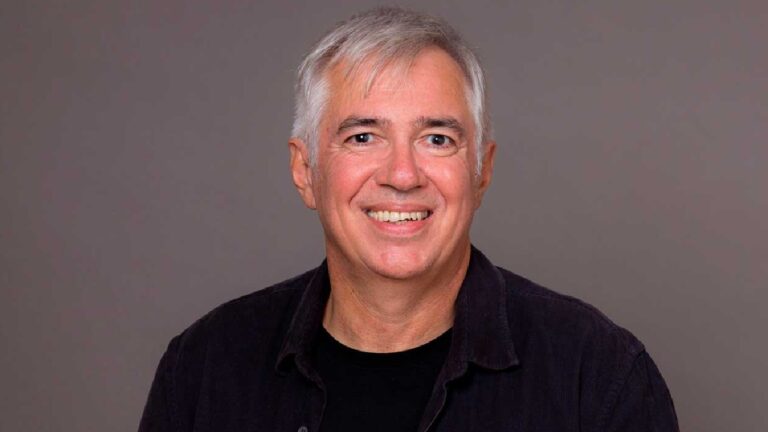 A imagem mostra um homem sorridente, de cabelos grisalhos curtos, vestindo uma camisa escura sobre uma camiseta preta, posando contra um fundo neutro. A expressão é simpática e transmite naturalidade