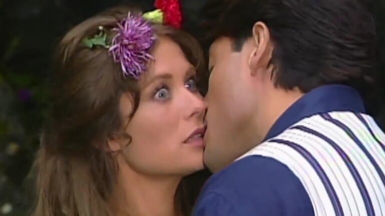 Mulher com flores no cabelo parece surpresa enquanto um homem se aproxima para beijá-la, em uma cena dramática típica de novela ou série romântica