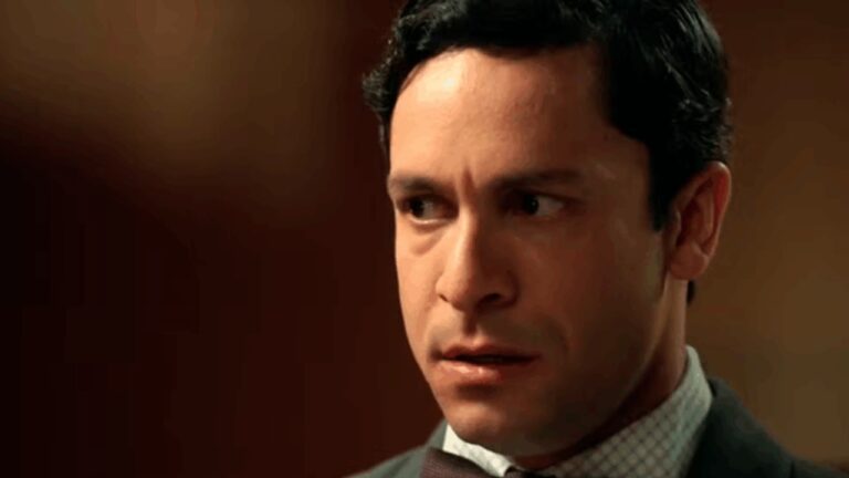 Homem de expressão tensa e olhar preocupado aparece em close-up, vestindo camisa social e paletó, em ambiente com iluminação quente e fundo desfocado