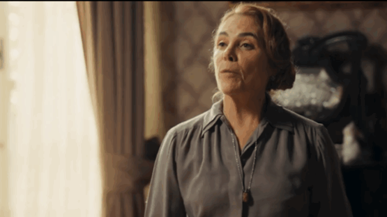 Mulher de expressão séria está em um ambiente interno com cortinas claras e papel de parede antigo, vestindo blusa cinza e colar simples, transmitindo tom de tensão ou reflexão