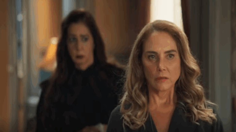 Cena mostra duas mulheres em um ambiente interno bem iluminado. A mulher em primeiro plano, de cabelos loiros ondulados, tem expressão séria e olhar fixo à frente, enquanto a mulher ao fundo, de cabelos castanhos escuros, parece surpresa ou preocupada