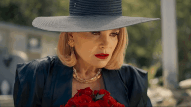 Mulher elegante de chapéu largo azul e batom vermelho segura um buquê de rosas vermelhas, com expressão séria e olhar intenso. A cena ocorre ao ar livre, sob luz natural, transmitindo um clima de mistério e sofisticação