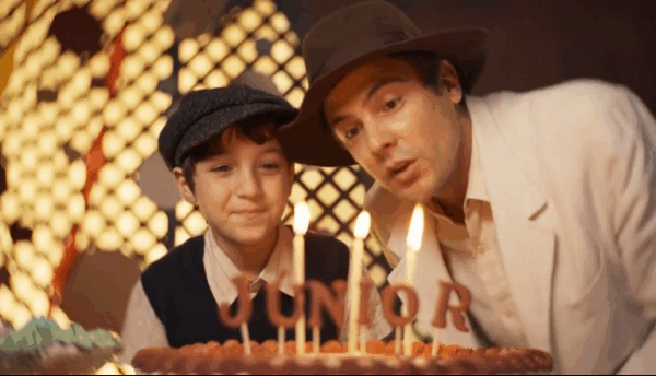 Criança e homem com chapéus observam bolo de aniversário com velas acesas e nome Junior em cena festiva