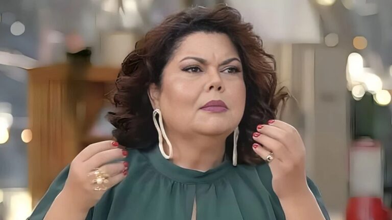 Mulher de expressão séria ajusta os cabelos com as mãos, exibindo unhas vermelhas e anéis chamativos. Ela usa vestido verde com detalhe no decote e brincos longos, posando em ambiente interno desfocado ao fundo