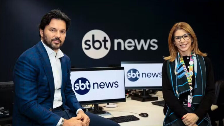 Fabio Faria de terno azul e Daniela Beyruti de óculos e lenço, posando em frente ao logo SBT News na redação