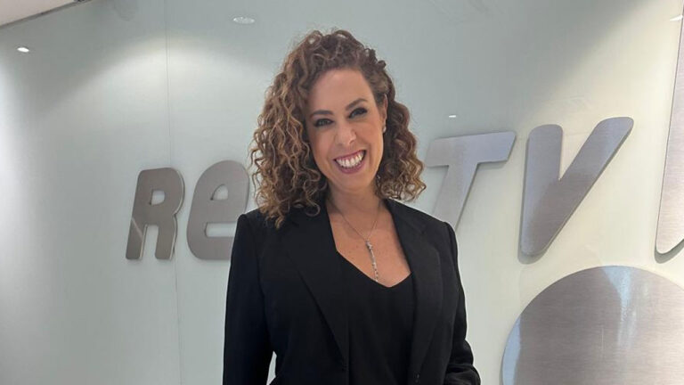 Fabíola Cidral sorrindo, com cabelo cacheado e blazer preto, em frente ao logo da RedeTV! na sede da emissora