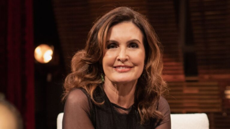 Mulher de cabelos castanhos e vestido preto sorri durante gravação em estúdio com iluminação suave e fundo amadeirado